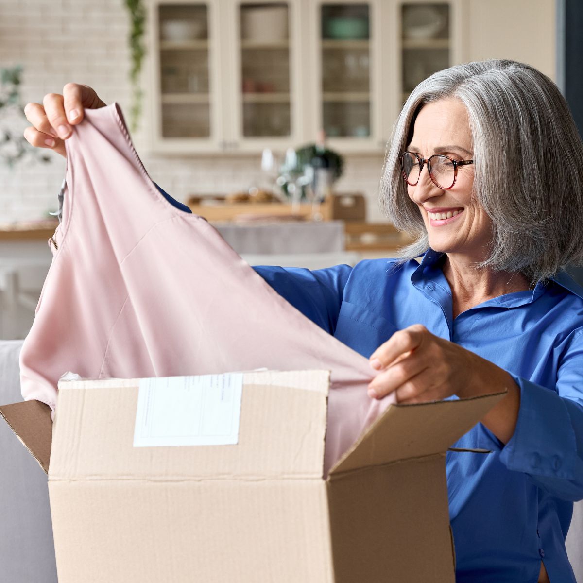 Happy-senior-middle-60s-aged-woman-opening-box-with-ordered-clothes-at-home-on-couch